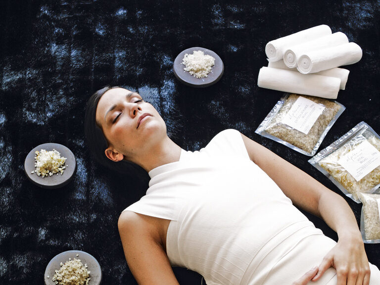 Eine Frau in einem weißen Kleid liegt auf dem Boden umgeben von zahlreichen verschiedenen Salzen der Marke Alpienne, die als Massage Pflegeprodukte verwendet werden können.