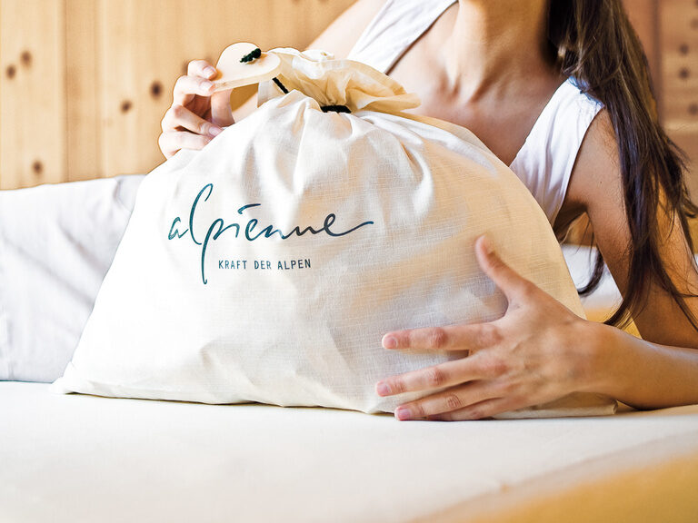 Eine Frau sitzt auf einem Bett in einem gemütlichen Schlafzimmer und präsentiert eine Tasche mit Kosmetikprodukten von der Marke Alpienne.