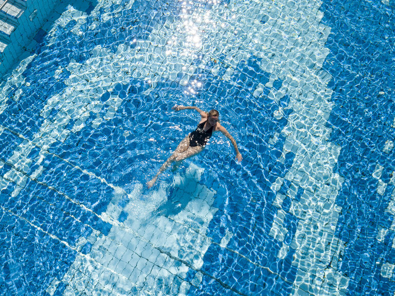 Ein Hotelgast schwimmt erholt im Aussenpool des Wellnesshotels Mooshof in Bodenmais umgeben von dem sonnenbeschienenen Wasser.