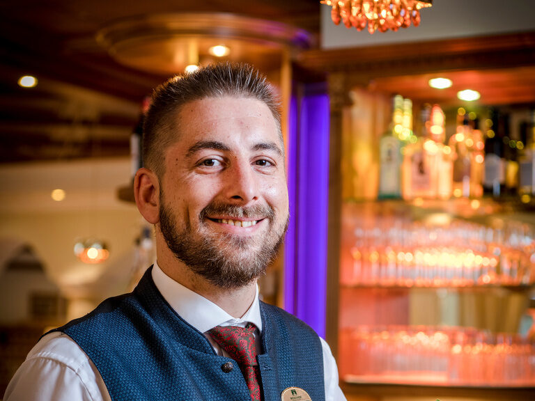 Der Barkeeper des Hotels Mooshof steht mit einem Cocktail in der Hand unter den gemütlichen Lichtern des Bartresens.