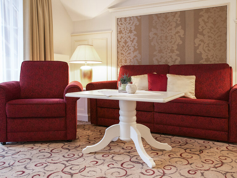 Die weiche rote Sitzgarnitur und ein Tisch im Wohnzimmer der Berkhain Suite Deluxe des Hotels Mooshof in Bayern laden zum entspannen ein.