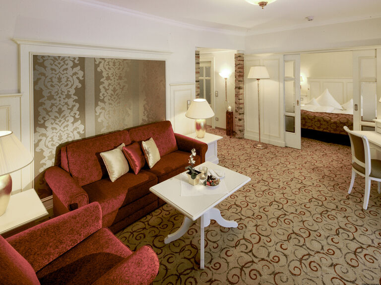 Gemütliches Wohnzimmerdesign in der Birkenhain-Suite des 4-Sterne-Hotels Mooshof mit roten Sitzgelegenheiten und eleganten weißen Tischen.