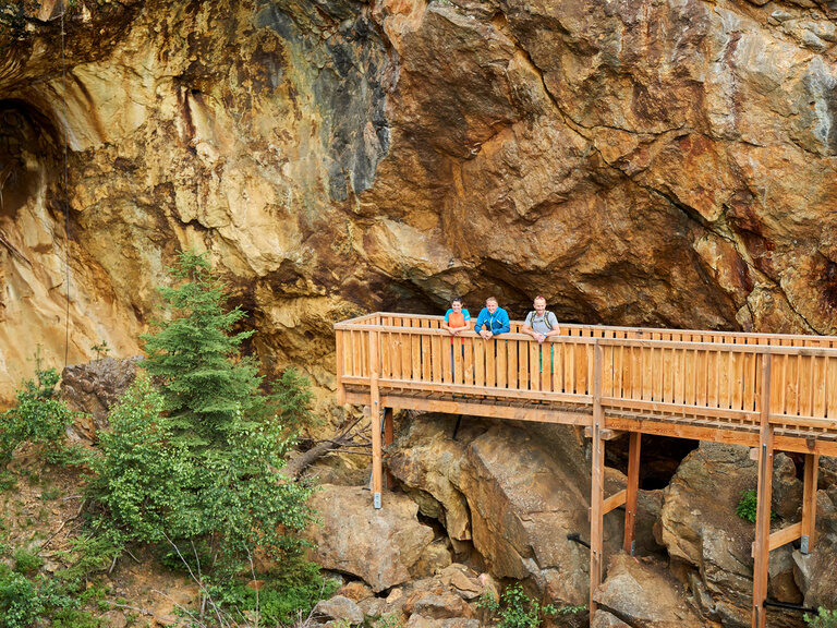 Eine Gruppe pausiert auf einer Holzbrücke vor einer imposanten Felsklippe.