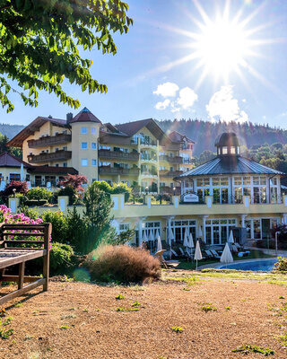 Eine einladende Bank unter einem Baum in dem blühenden Garten mit Blick auf das Wellnesshotel Mooshof in Bodenmais.