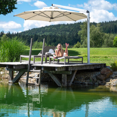 Eine Frau verweilt entspannt unter einem schattenspendenden Sonnenschirm auf dem Steg des Naturbadesees im Wellnesshotel Mooshof im Bayerischen Wald.