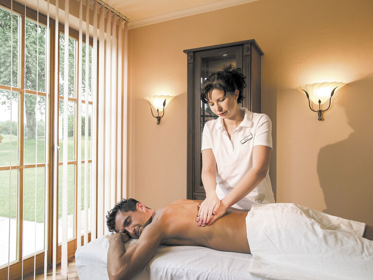 Ein Mann befindet sich auf einem Massagetisch in einem beleuchteten Raum nahe dem Fenster und erhält eine erholsame Rückenmassage im Wellnesshotel Mooshof im Bayerischen Wald.