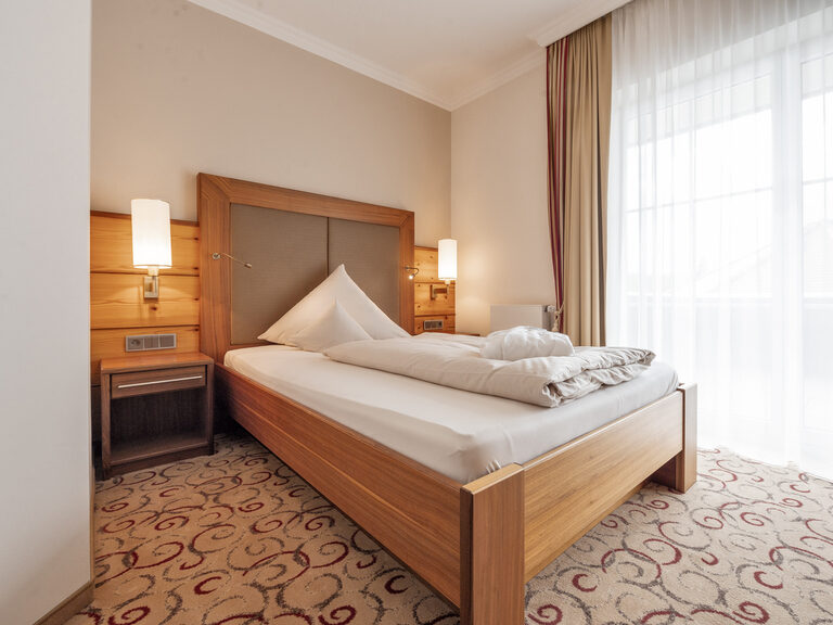 Das komfortable Hotelzimmer des Wellnesshotels Mooshof im Bayerischen Wald im Erkersuite-Stil mit einem gemütlichen Bett und beleuchteten Nachtischen.
