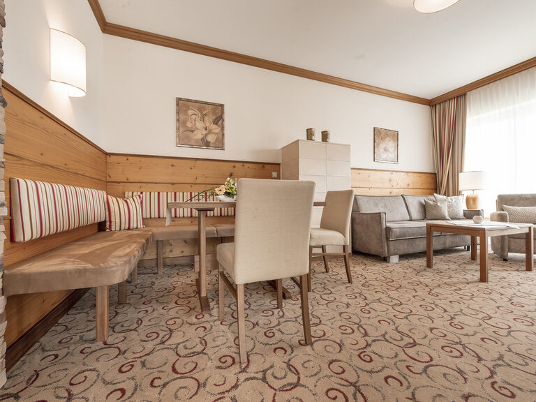Das geräumige Erkersuite-Wohnzimmer des Hotels Mooshof in Bodenmais mit stilvollen Holzmöbeln zum Sitzen und Panoramaausblick aus dem großen Fenster.