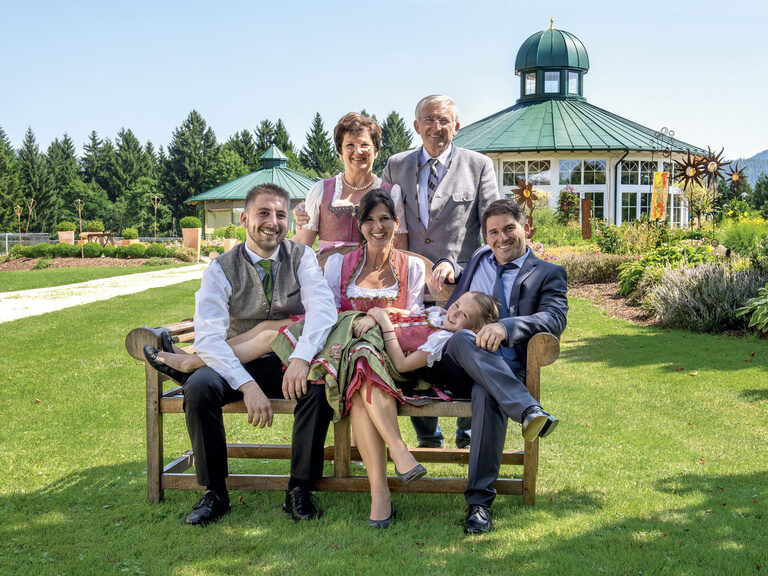 Die Gastgeber Familie Holzer sitzt entspannt auf einer Bank im blühenden Garten mit Pavillion des Wellnesshotels Mooshof in Bayern.