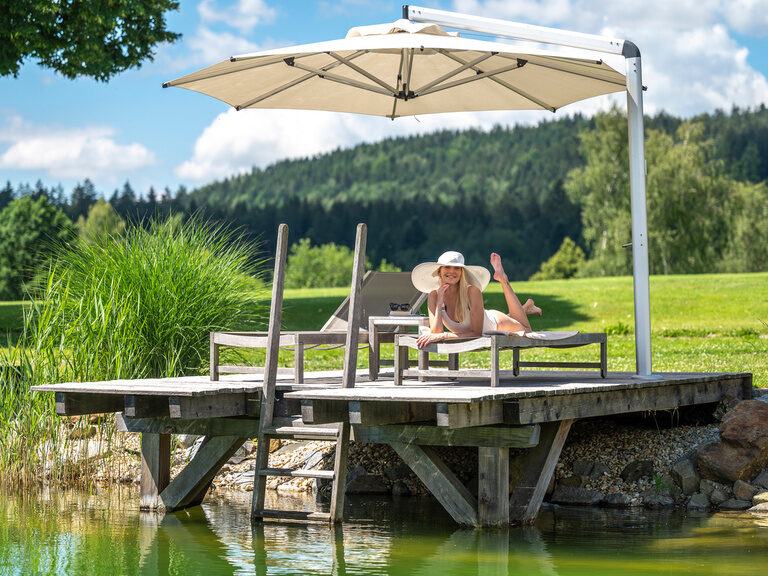 Die Urlauberin genießt die Ruhe auf dem Steg des Mooshof-Resorts unter einem schattenspendenden Schirm.