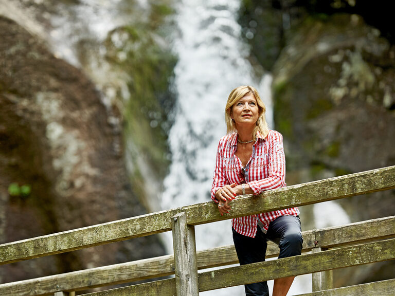 Eine Frau blick von einer Brücke mit Wasserfall im Hintergrund in die Ferne.