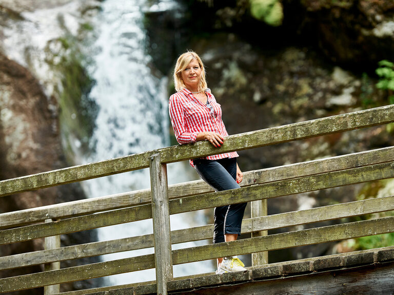 Eine Frau blickt von einer Brücke mit Wasserfall im Hintergrund nach unten.