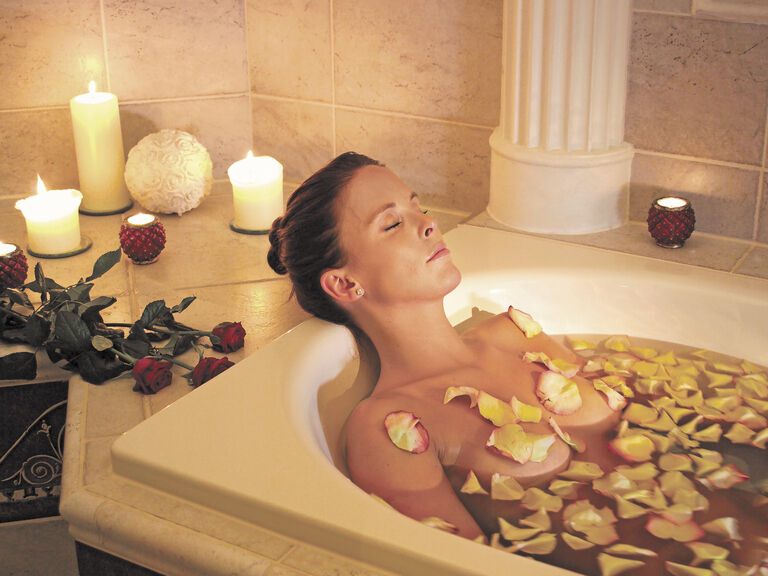 Eine Frau genießt ein Bad mit Rosenblütenblättern in einer eleganten Badewanne, umgeben von Kerzenlicht im Wellnesshotel Mooshof im bayerischen Wald.