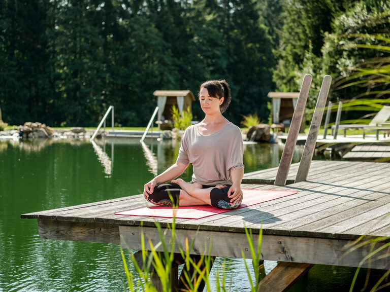 Eine Frau sitzt auf einem Steg und macht beruhigendes Yoga im Wellness & SPA Resort Mooshof.
