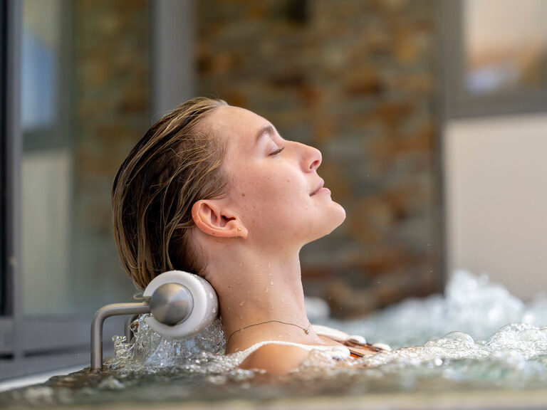 Eine Frau lehnt entspannt am Beckenrand und genießt das sprudelnde Wasser des Wellnessbereiches des Wellnesshotels Mooshof im Bayerischen Wald.