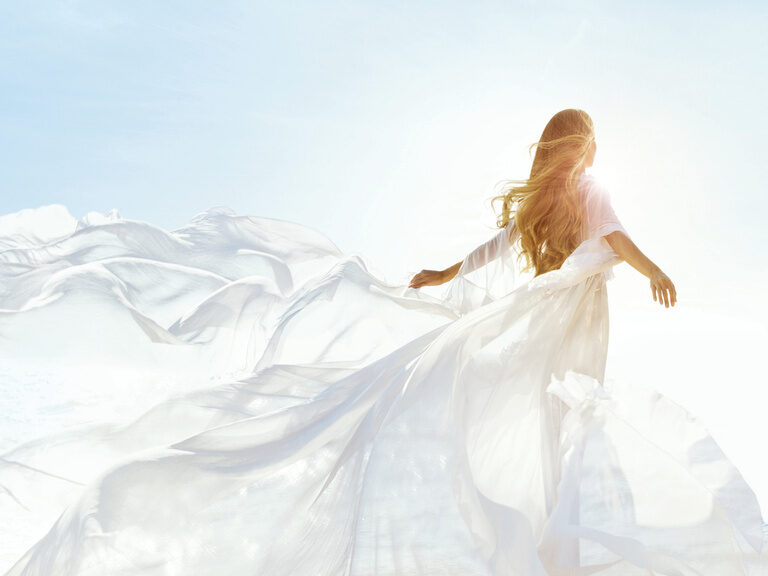 Ein weißes Kleid umspielt eine Frau, während sie mit Blick in die Sonne die Freiheit genießt.