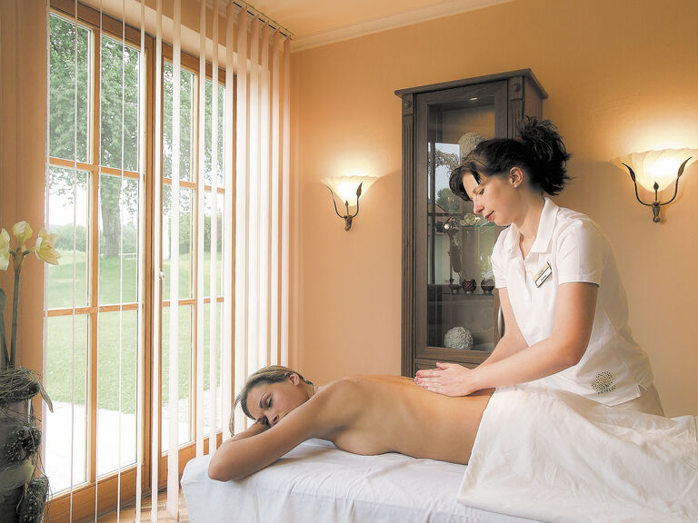 Eine Frau befindet sich auf einem Massagetisch in einem beleuchteten Raum nahe dem Fenster und erhält eine erholsame Rückenmassage im Wellnesshotel Mooshof in Bodenmais.