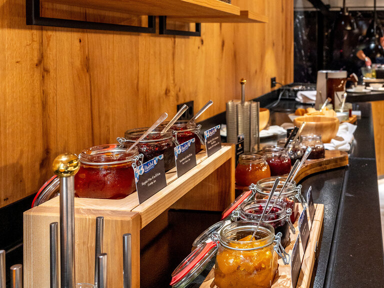 Eine hölzerne Theke im Frühstücksbuffetbereich des Wellnesshotels Mooshof in Bayern mit verschiedenen Frühstückszutaten in Gläsern.