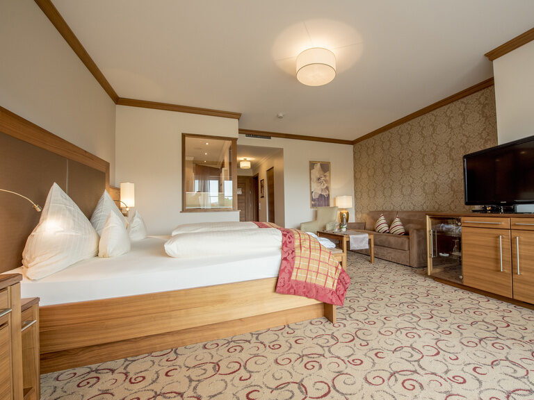 Das Gartensuite Schwalbennest Zimmer des Welllnesshotels Mooshof in Bodenmais mit einem großen Bett und einem, an die Wand montierten, Fernseher zur Unterhaltung.