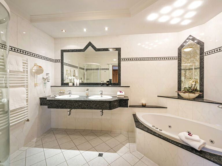Das hell beleuchtete Badezimmer in der Harlachbergsuite des Wellnesshotels Mooshof mit kontrastierenden schwarz-weißen Fliesen bei der Badewanne und dem Waschbecken.