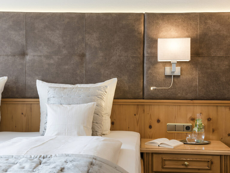 Ein rustikaler Holznachttisch neben dem kuscheligen Bett in der Juniorsuite Classic des Wellnesshotels Mooshof im Bayerischen Wald..