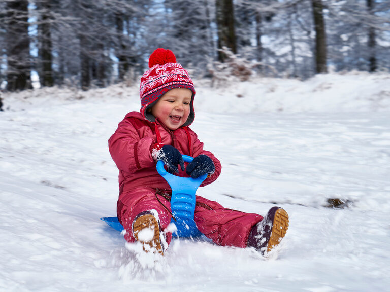 Ein lächelndes Kind fährt auf einem blauen Schlitten einen Hügel hinunter.
