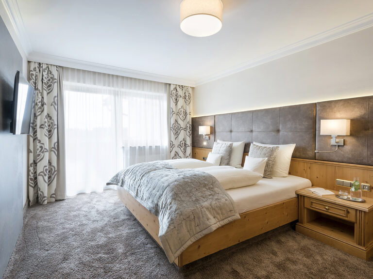 Stilvoll eingerichtetes Schlafzimmer in der Komfortsuite des Wellnesshotels Mooshof in Bayern mit einem bequemen Bett und Fernseher an der Wand..