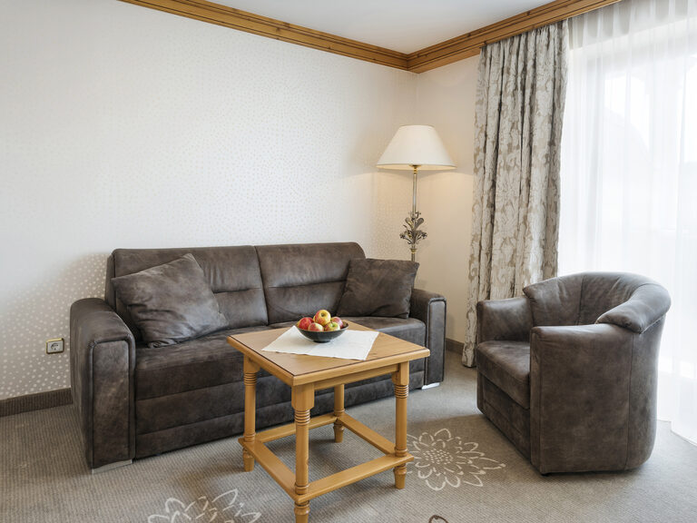 Einladender Wohnbereich im Landhaussuiten Stil des Wellnesshotels Mooshof im Bayerischen Wald mit einer Couch und einem Sessel und einem eleganten Couchtisch mit Obstschale in der Mitte.