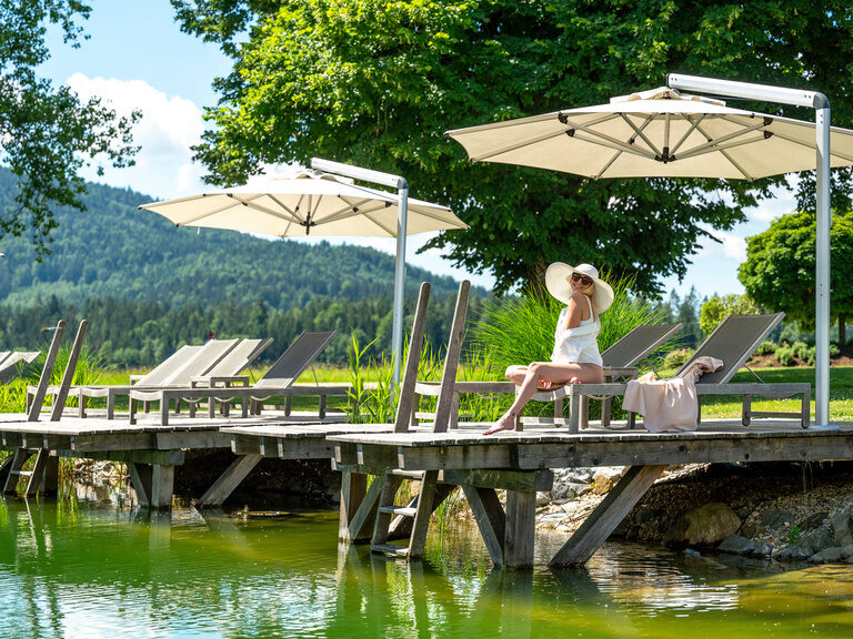 Eine Frau im Schatten eines Sonnenschirms am Naturbadesee des Hotel Mooshof.