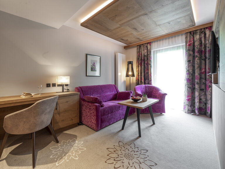 Der Loungebereich des Zimmers Typ 1 im Wellnesshotel Mooshof im Bayerischen Wald mit gemütlichem lila Sofa und Flachbildfernseher.
