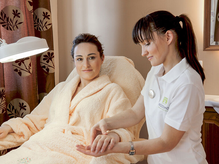 Eine Kosmetikerin bietet persönliche Nagelpflege in dem entspannenden Umfeld des Vitalresorts im Hotel Mooshof an, während die Frau lächelt.