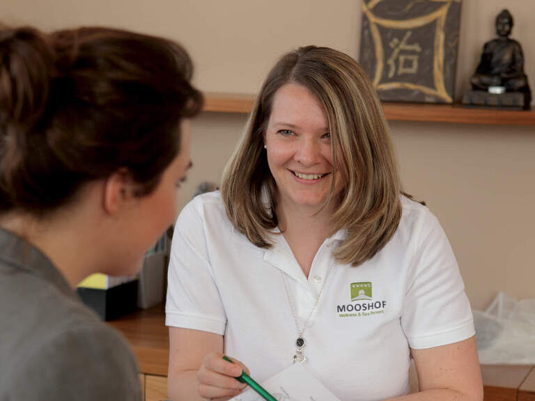 Eine Heilpraktikerin in Berufskleidung des Wellnesshotels Mooshof im Bayerischen Wald berät eine Frau für eine Behandlung in der Naturheilkundlichen Praxis.
