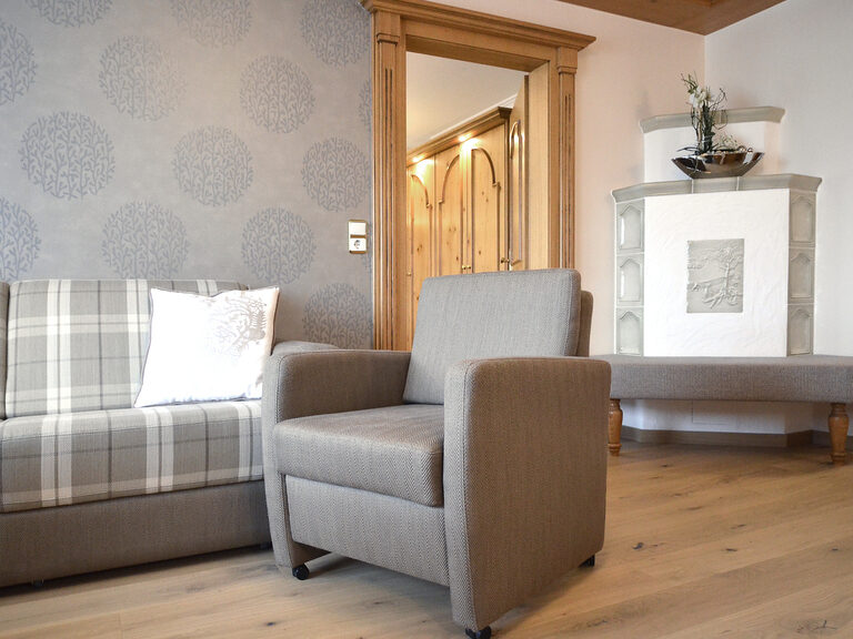 Die Komfortsuite im Wellnesshotel Mooshof in Bodenmais bietet eine stilvolle Couch und einen Sessel vor einem gemütlichen Kachelofen im Wohnbereich.