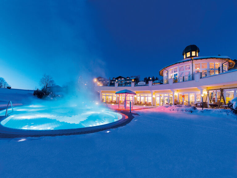 Das Wellnesshotel Mooshof in Bodenmais mit dem heißen Pool im Vordergrund, umgeben von Abenddämmerung im winterlichen Bayerischen Wald.