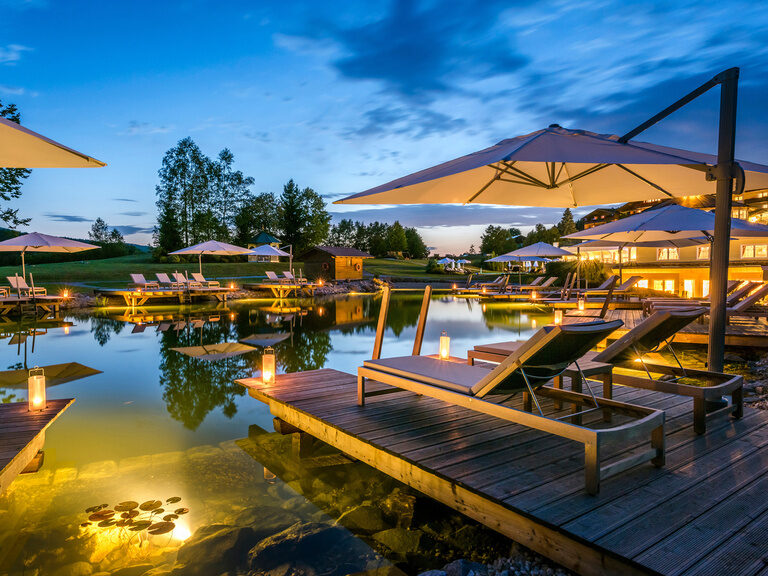 Der hölzerne Steg mit bequemen Liegen und Sonnenschirmen am Naturbadesee des Wellnesshotels Mooshof in Bayern.