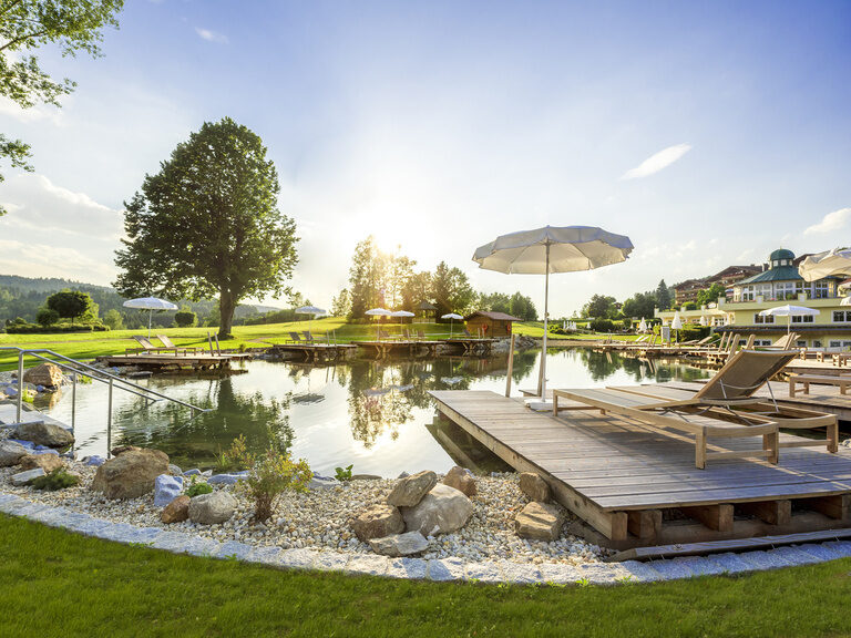 Malerischer Naturbadesee im Wellness & SPA Resort Mooshof im Bayerischen Wald mit umliegenden Sonnenschirmen und Liegen im grünen Garten bei Sonnenuntergang.
