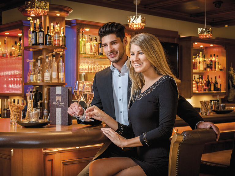 Gemütliche Bar im Hotel Mooshof im Bayerischen Wald mit zwei Gästen in eleganter Kleidung, die ein Glas Wein genießen..