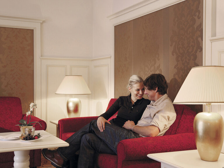 Ein Paar verbringt seine gemeinsame Zeit glücklich auf einem roten Sofa im Wohnzimmer der Birkenhain-Suite im Wellnesshotel Mooshof im Bayerischen Wald..