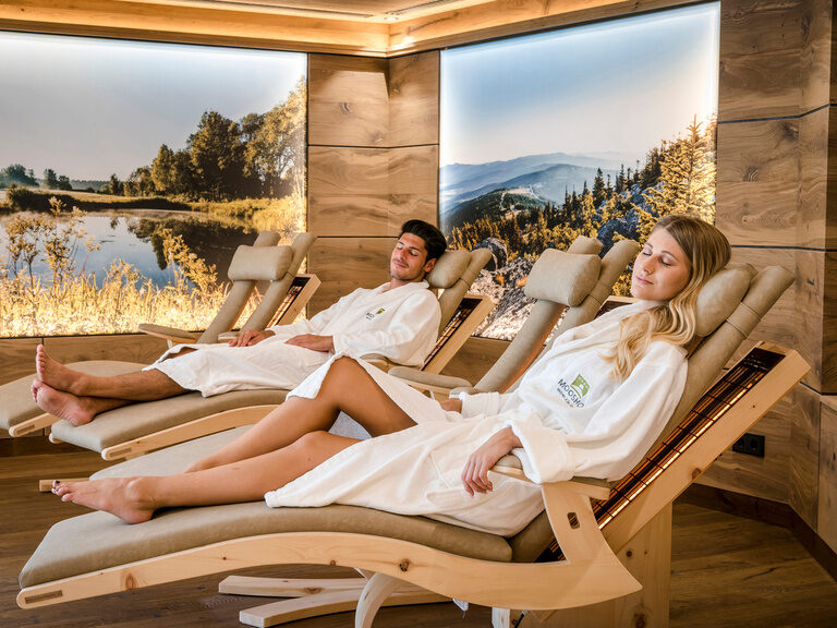 Ein Paar erholt sich in einem Ruheraum des Wellnesshotels Mooshof auf Ruheliegen, umgeben von einer harmonischen Beleuchtung und Bildern der Urlaubsumgebung.