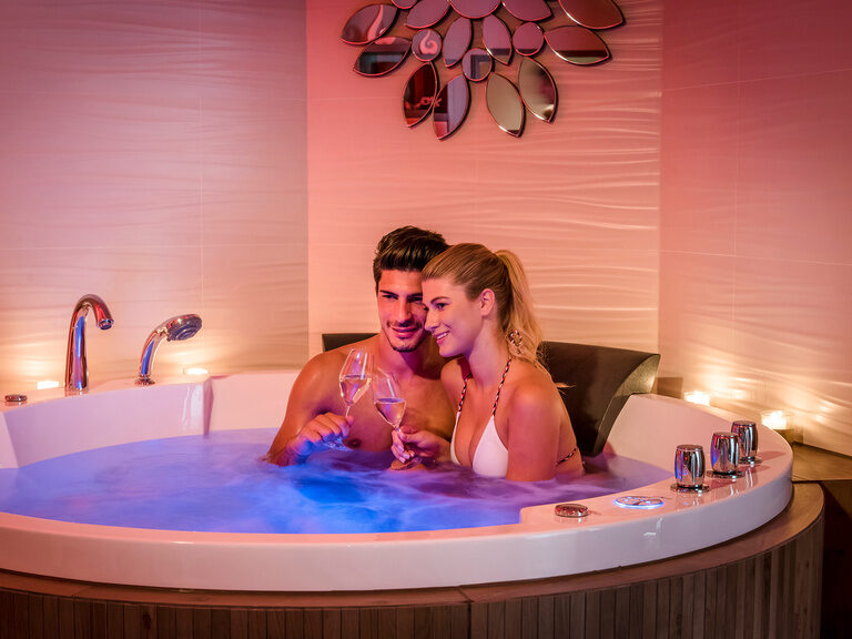 Kerzen schaffen ein romantisches Ambiente für ein Paar im blau beleuchteten Whirlpool in der private Suite des Wellnesshotels Mooshof im Bayerischen Wald.
