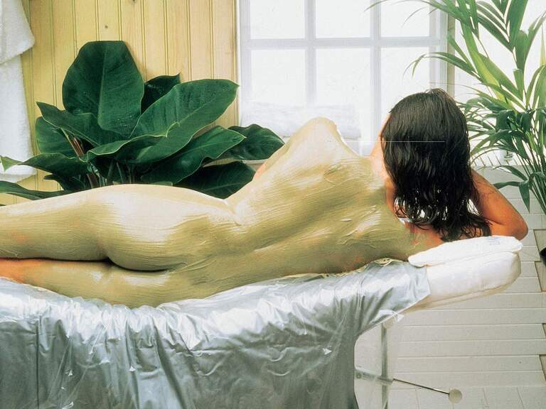 Gemütlicher Wellnessbereich im Wellnesshotel Mooshof mit einer Pflanze neben dem Massagetisch, auf dem eine Frau mit Ganzkörpermaske liegt.