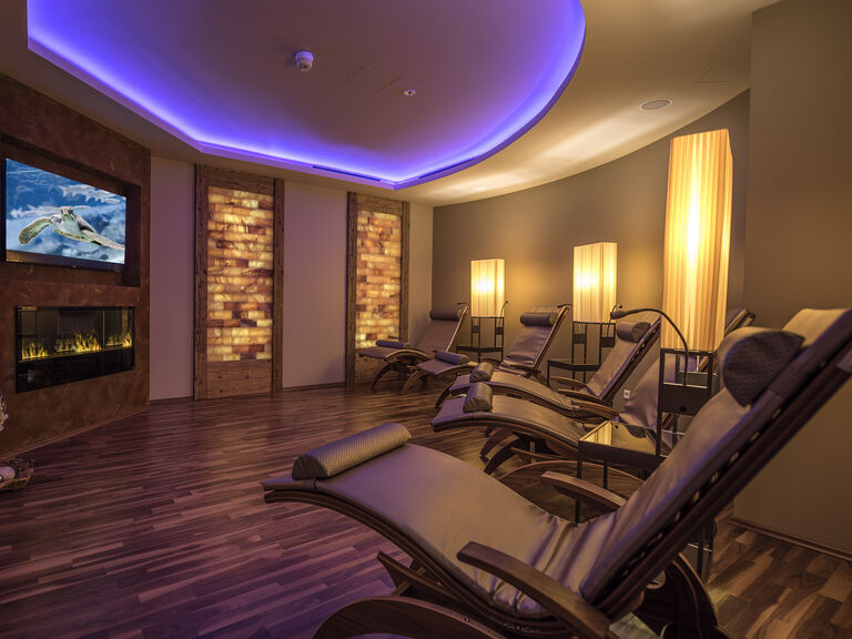 Der gemütliche Salzsteinruheraum mit Kamin, Liegen und einem Flachbildfernseher, perfekt für eine entspannte Auszeit im Wellnesshotel Mooshof in Bodenmais.