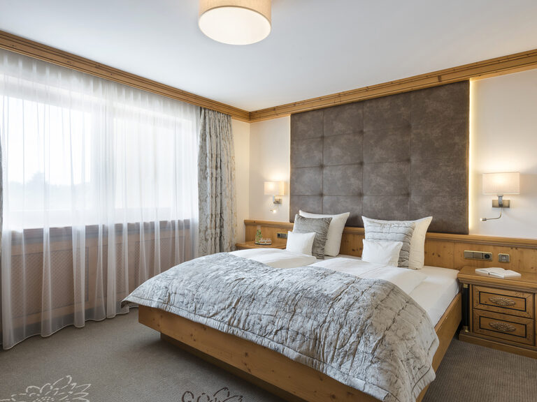 Gemütliches Bett im Schlafzimmer der Landhaussuiten des Wellnesshotels Mooshof in Bayern neben einem lichtdurchfluteten Fenster.