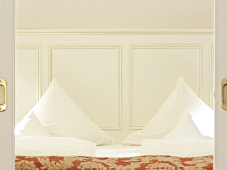 Das weiße Bett in der Birkenhain-Suite des Wellnesshotels Mooshof im Bayerischen Wald bietet einen Blick auf die geöffnete Zimmertür.