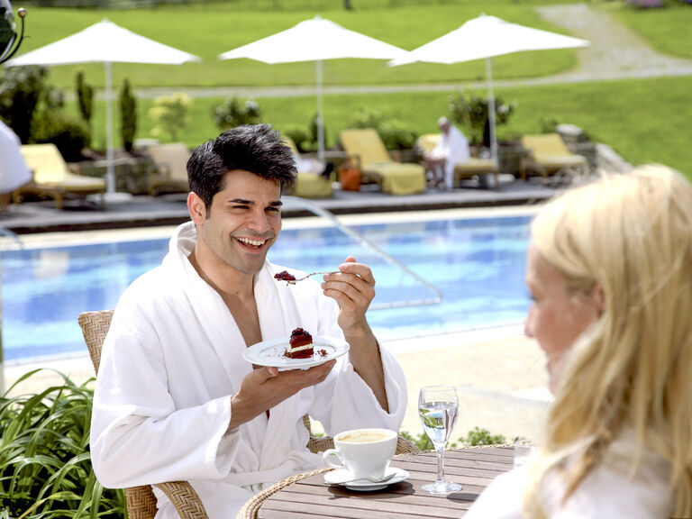Ein Paar genießt lachend Drinks und Kuchen am Spa Bistro in der Nähe des Aussenpools vom Wellnesshotel Mooshof in Bodenmais.