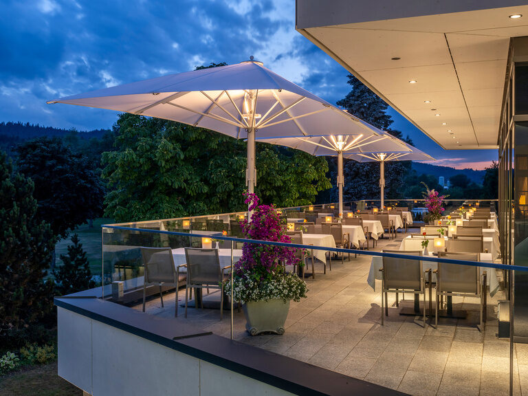 Die mit Stühlen und Sonnenschirmen ausgestattete Panoramaterrasse des Wellnesshotels Mooshof in Bodenmais bei stimmungsvoller Beleuchtung nachts.