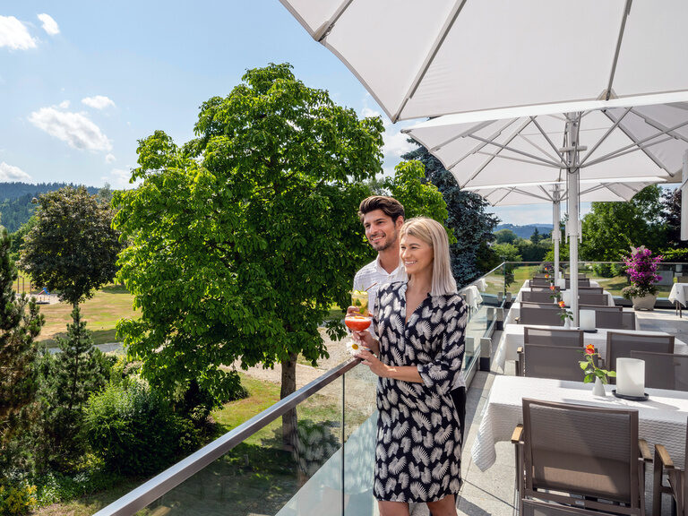 Ein Paar genießt gemeinsam mit einem alkoholischen Getränk seine Zeit auf der sonnenbeschienenen Terrasse des Wellnesshotels Mooshof.