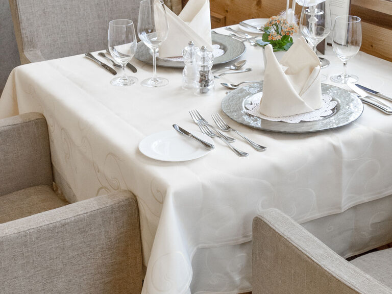 Geschmackvoll eingedeckter Tisch mit Gläsern und Tellern auf weißem Tischläufer im Speisesaal der Erkerlounge des Hotels Mooshof in Bayern.