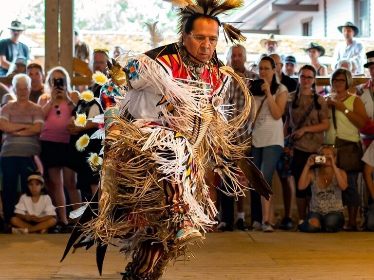Im Mittelpunkt steht ein Performer im Federindianerkostüm inmitten eines Schaulustigenpulks bei einer Kulturveranstaltung in Pullmancity.