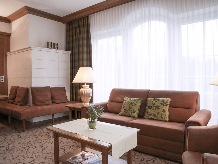 Geschmackvoll eingerichtetes Wohnzimmer in der Turmsuite Kronberg des Hotels Mooshof in Bodenmais mit einem Sofa und Sessel und einer gemütlichen Eckbank an dem Kachelofen.